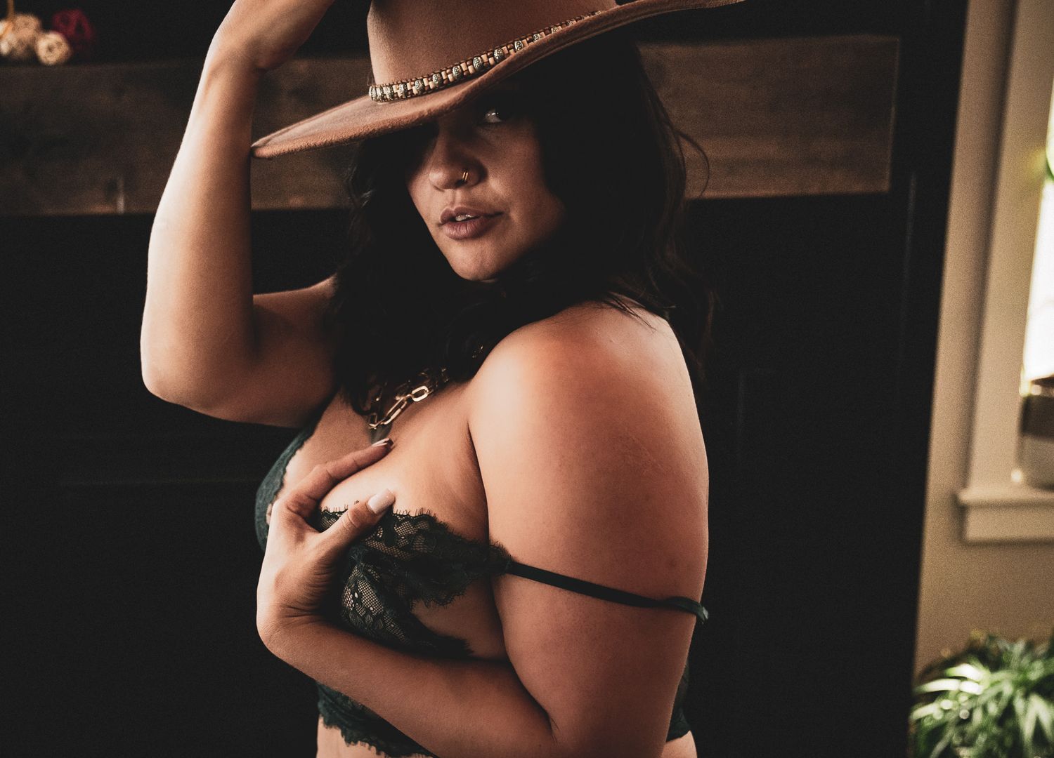 Curvy woman in black lingerie wearing cowboy hat posing in dimly lit room.