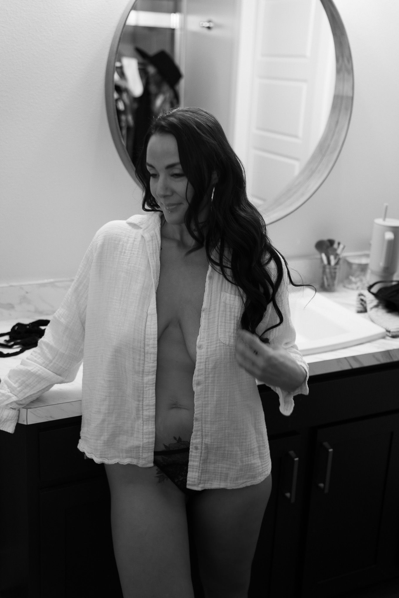 Black and white photo of brunette woman in open white shirt standing in bathroom by mirror.