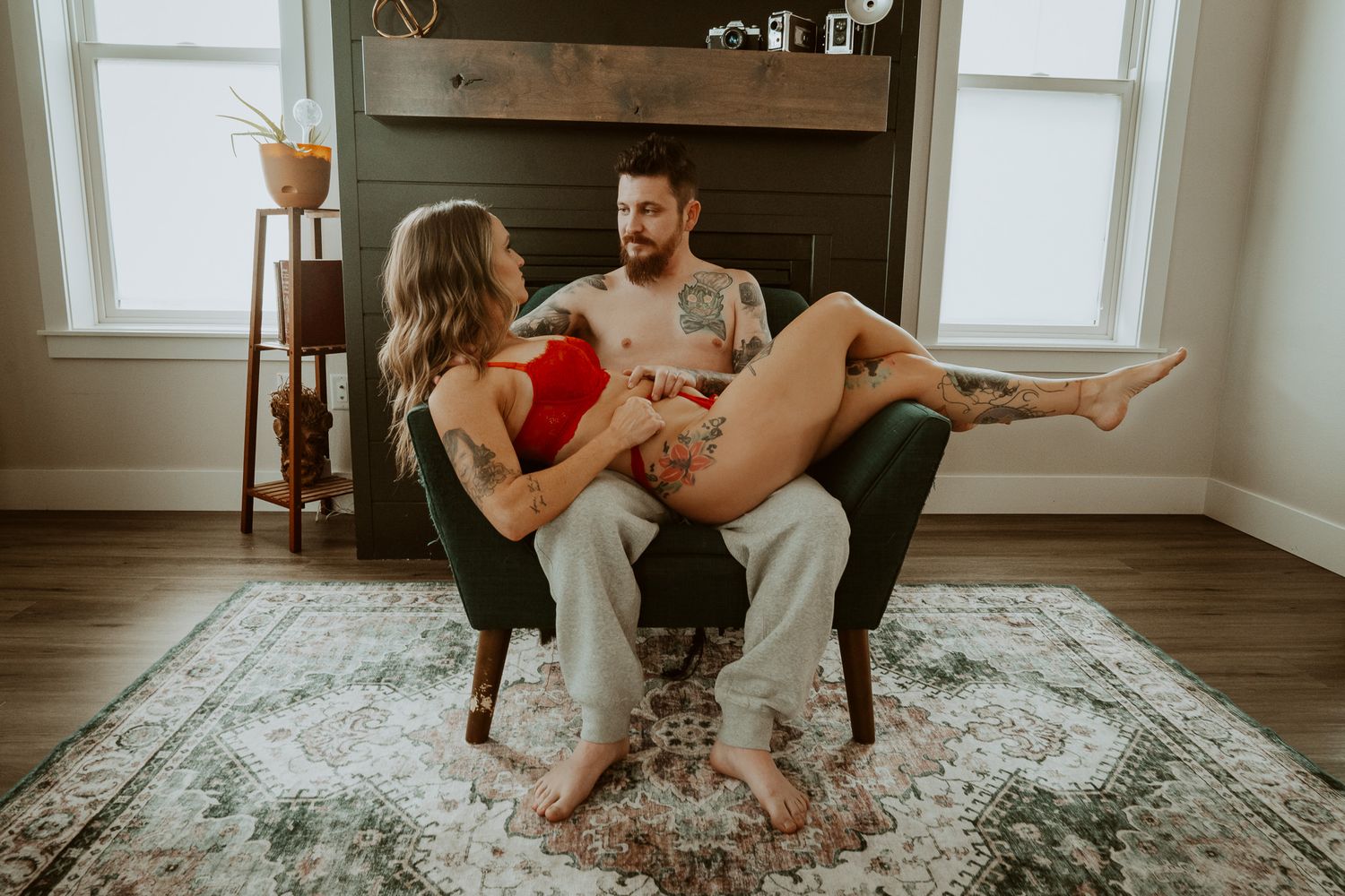 Tattooed couple sitting intimately in a chair together in a bright living room with hardwood floors.
