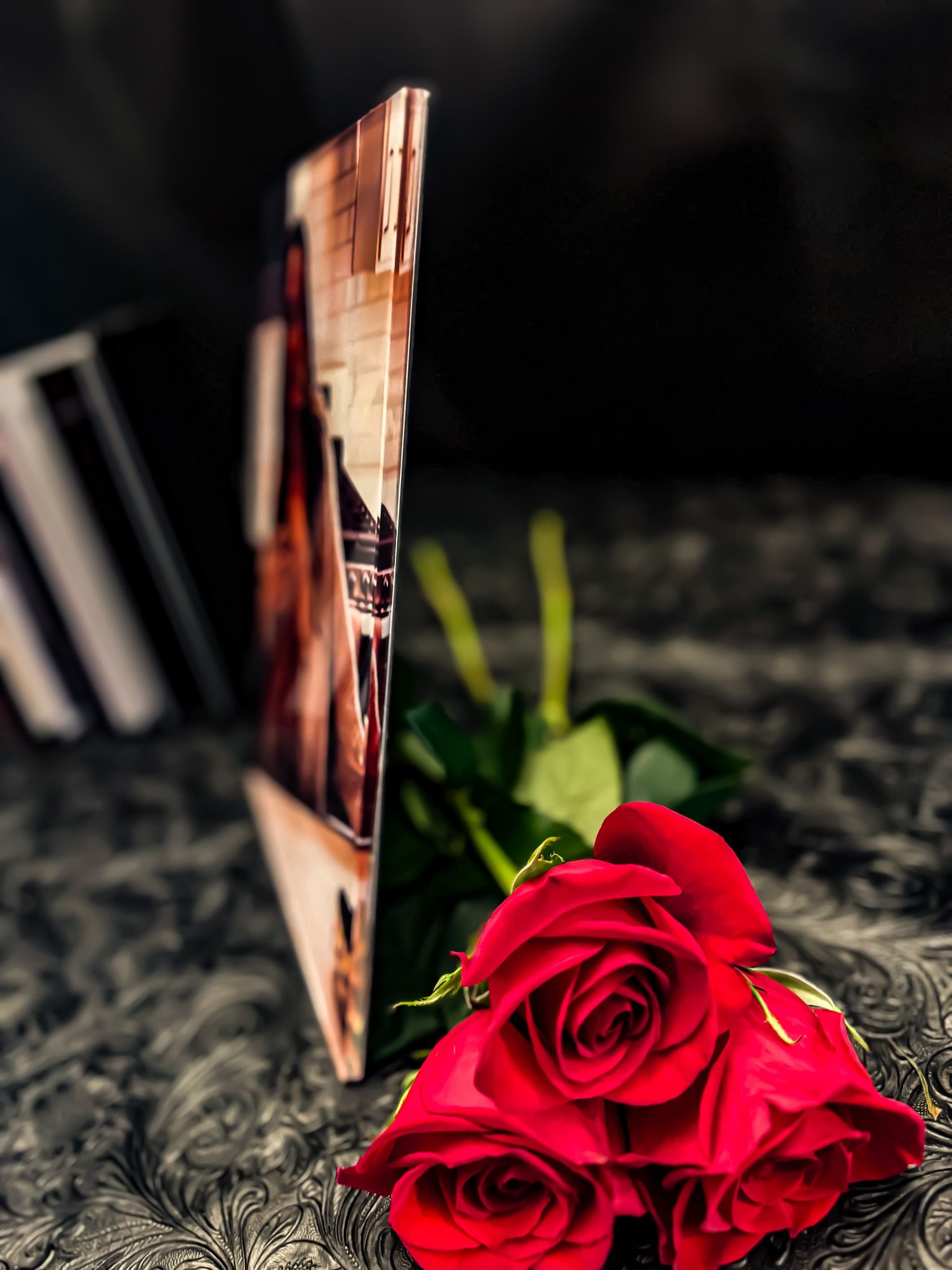 Red roses beside an open photo book standing upright on a dark embossed surface.