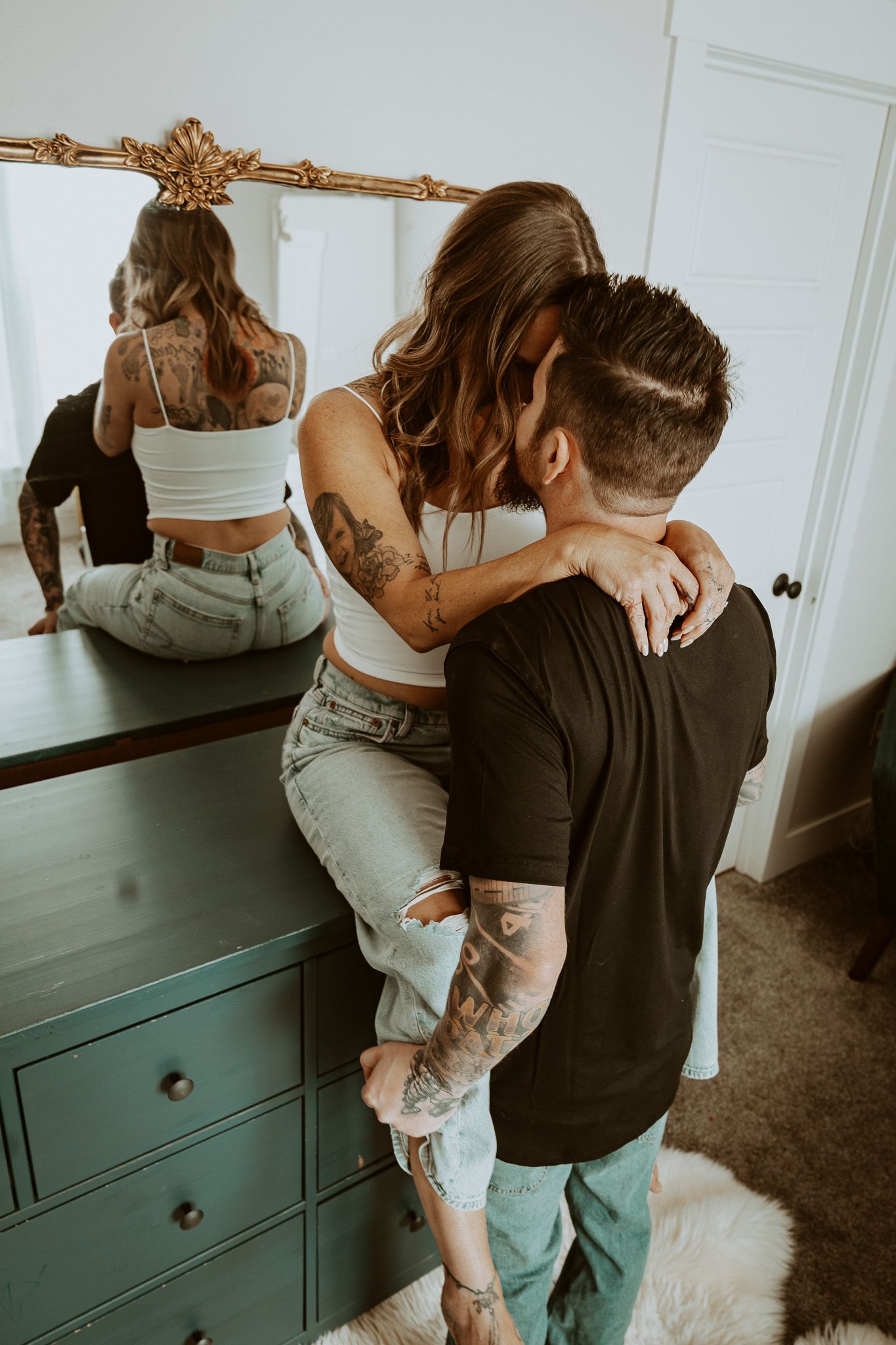 Tattooed couple embracing near a vintage mirror and teal dresser, romantic lifestyle photography session.
