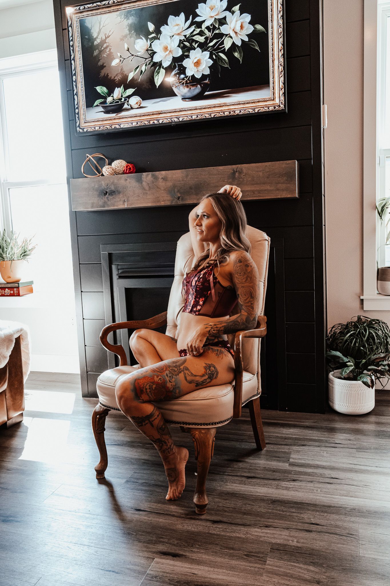 Heavily tattooed blonde woman in lingerie sitting in an ornate chair by a fireplace indoors.