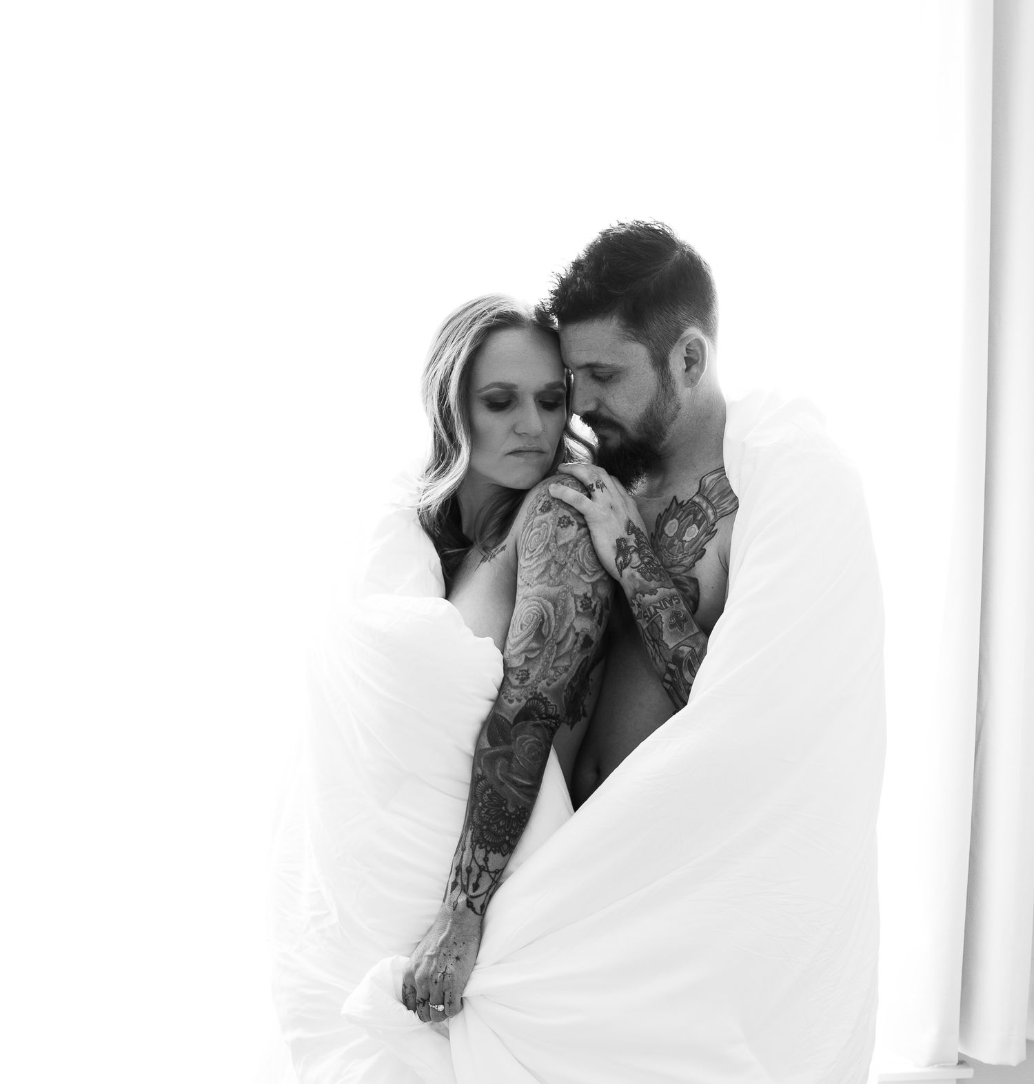 Black and white photo of a tattooed couple wrapped together in white sheets.
