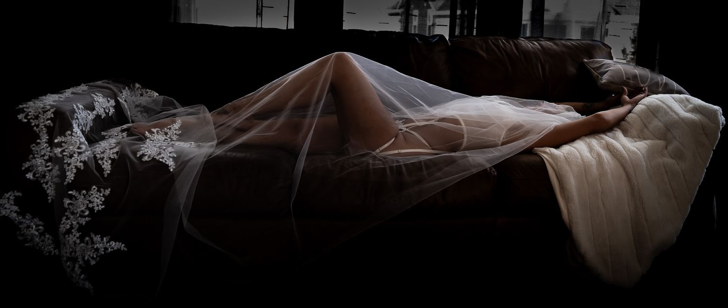 Artistic dark boudoir photo of woman draped in sheer lace bridal veil on bed.