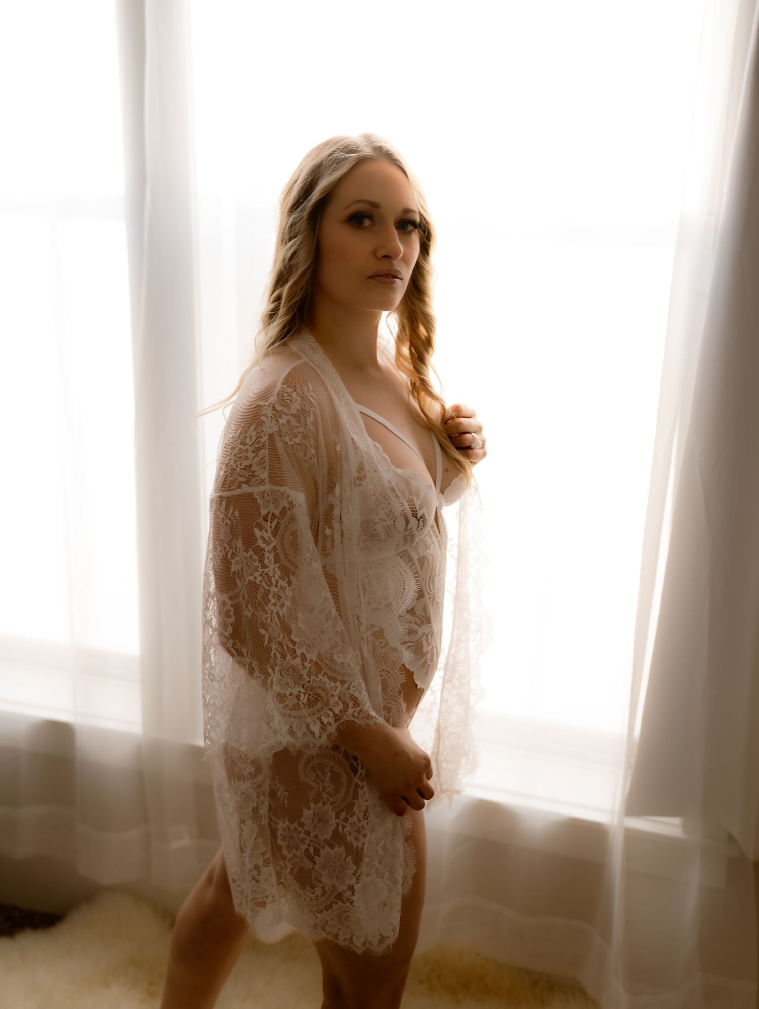 Blonde woman in sheer white lace robe standing before bright window in elegant boudoir photo.