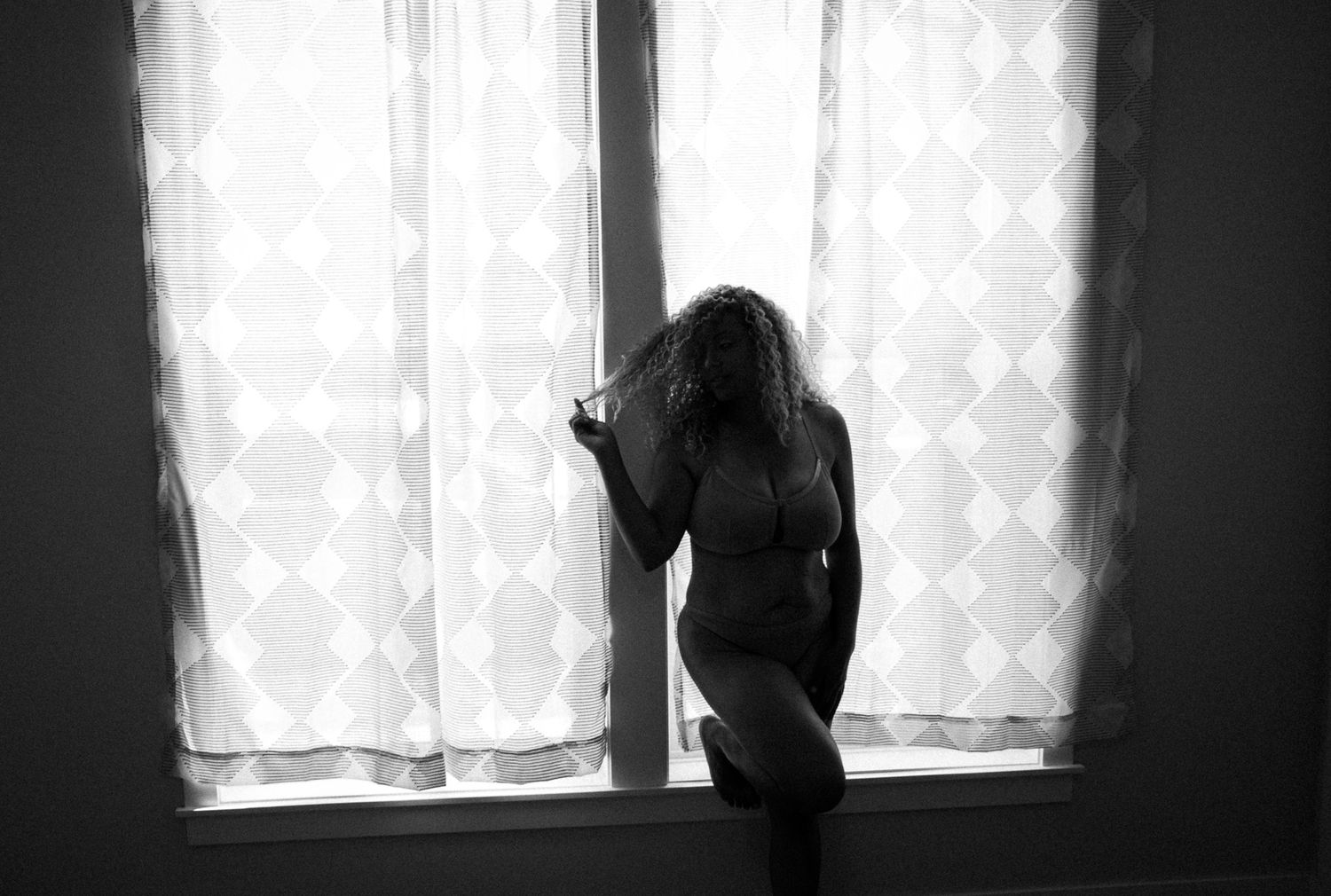 Black and white artistic silhouette of a curly-haired woman standing nude before a bright window.