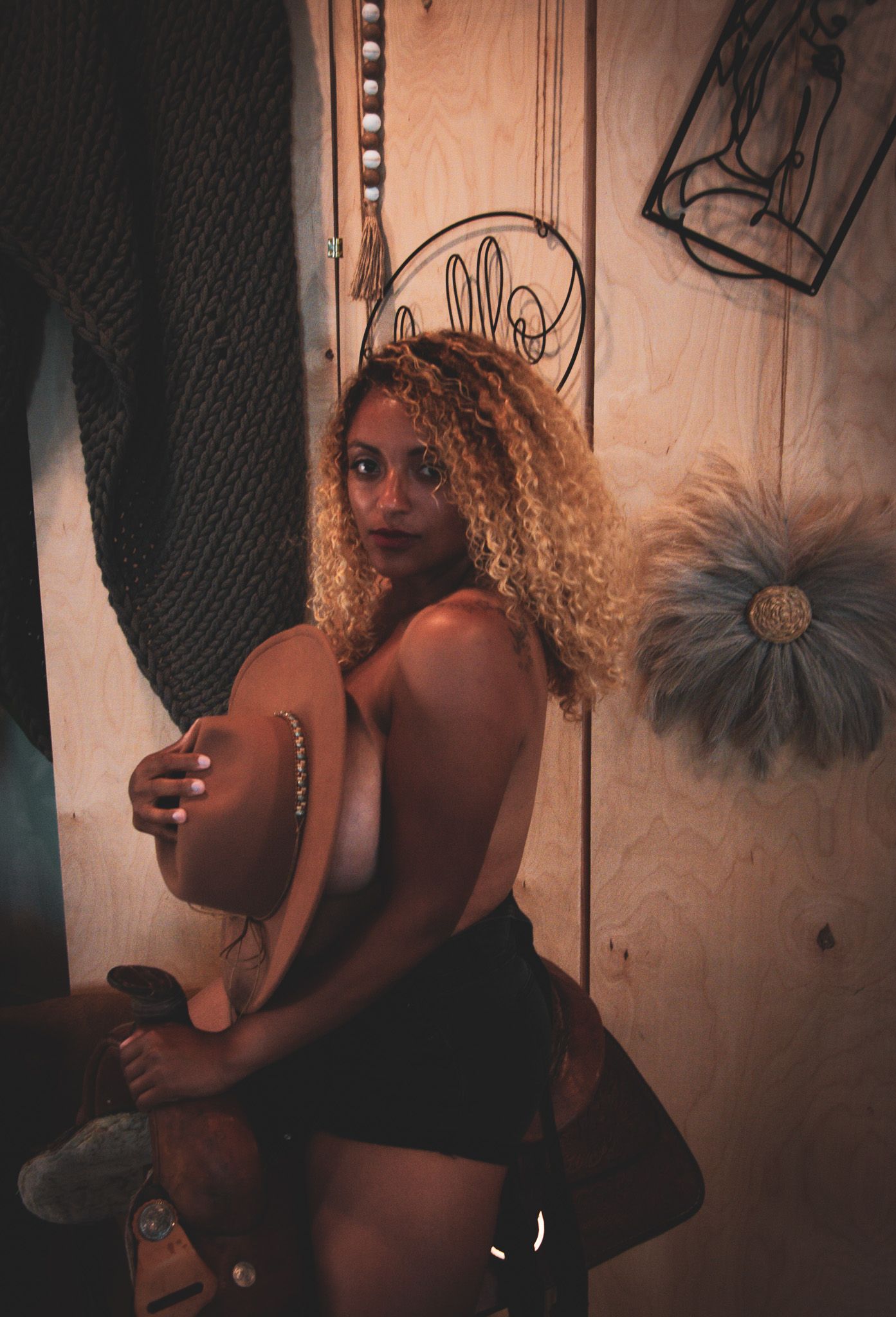 Woman with curly blonde hair posing topless holding a hat in a rustic wooden room interior.