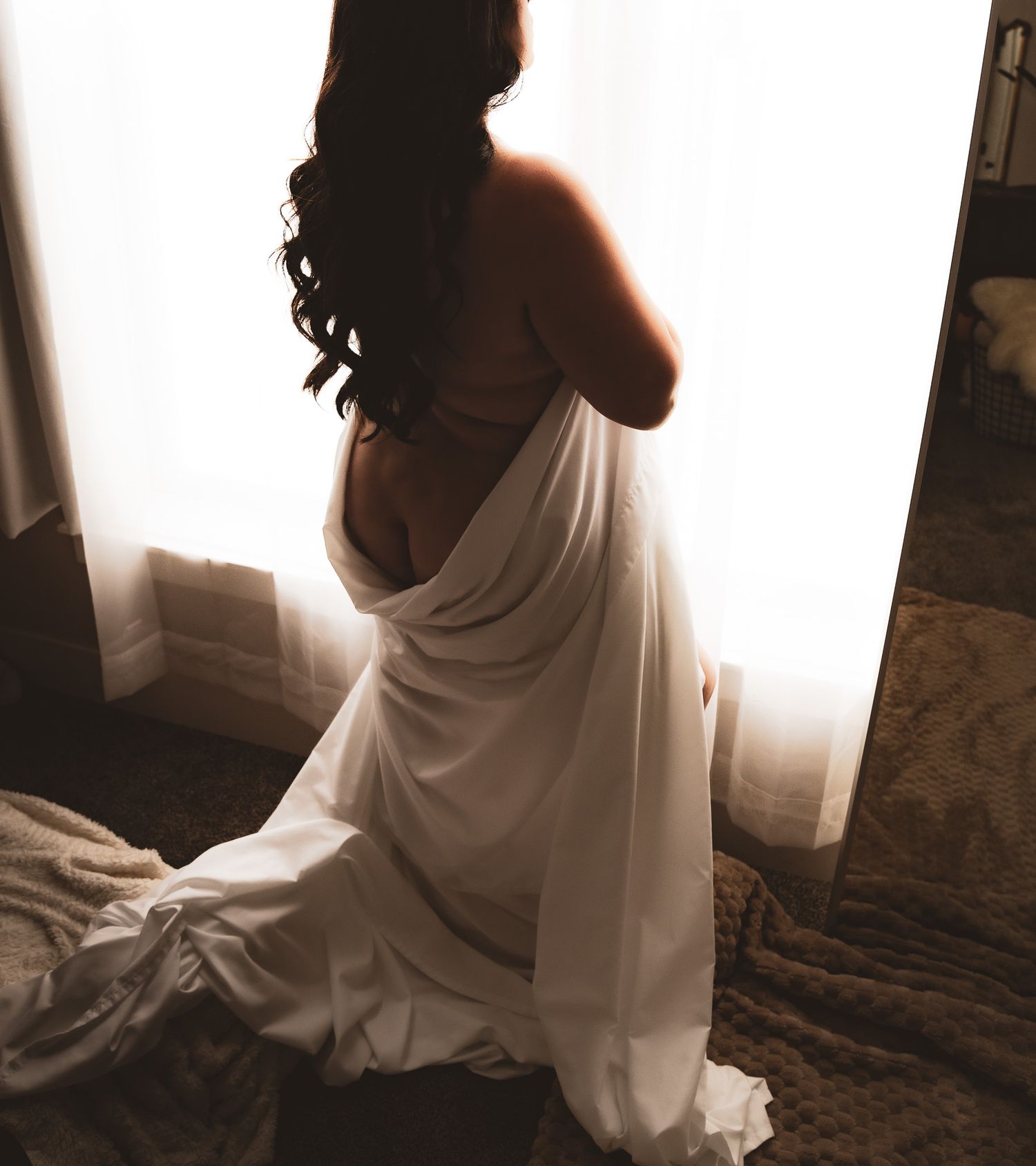A woman draped in white fabric stands backlit by a window in a bedroom.