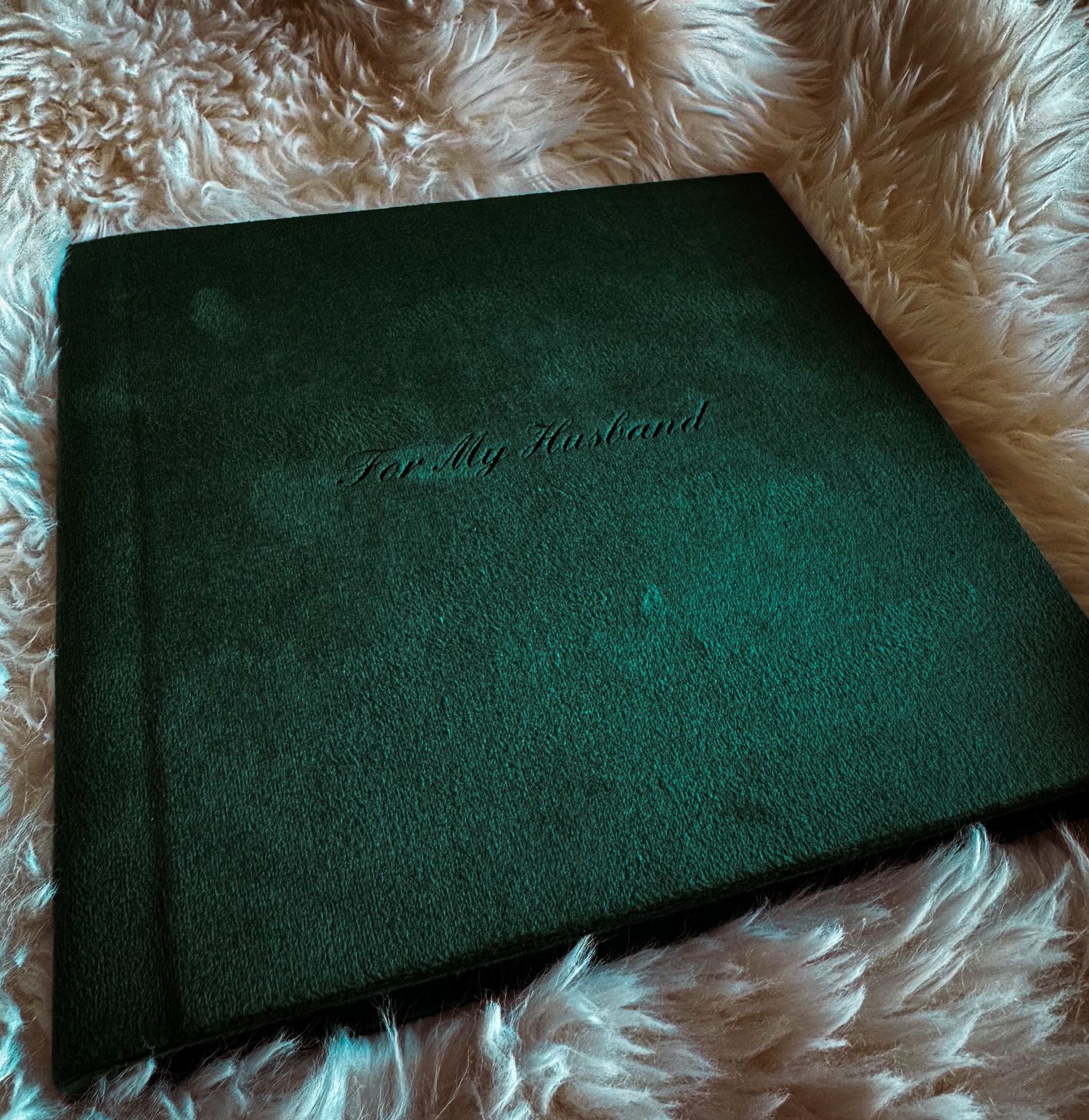 Dark green velvet photo album inscribed with For My Husband resting on a fluffy white fur surface.