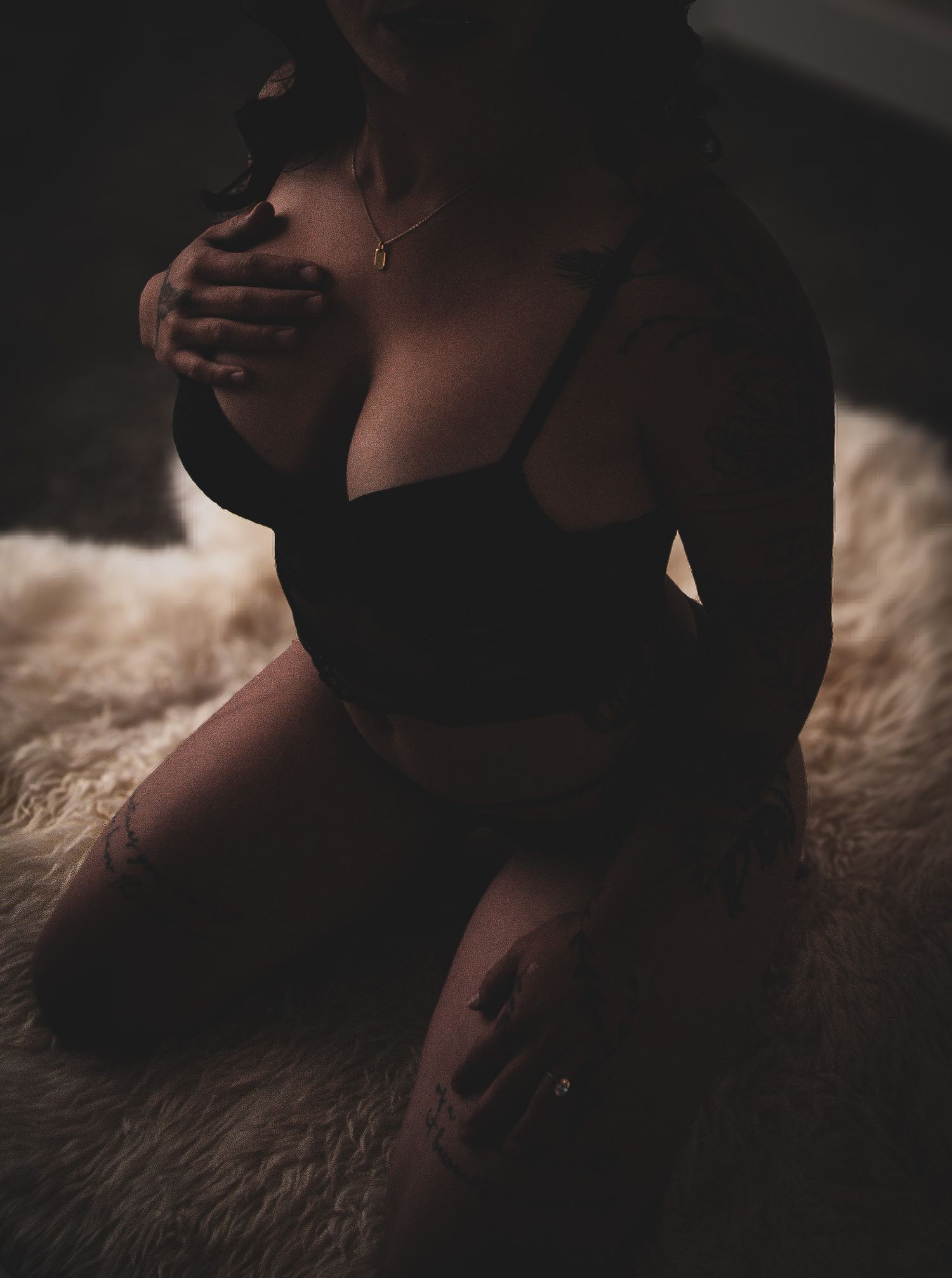 Woman in black lingerie kneeling on a fur rug with visible tattoos in moody dark lighting.