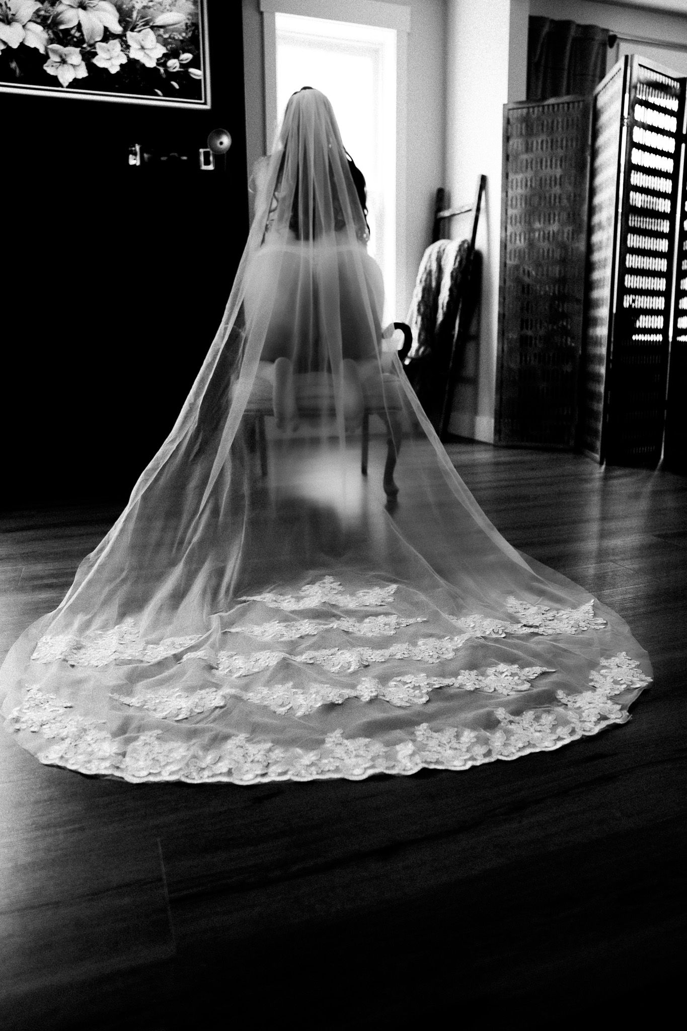 Black and white photo of a bride wearing a long lace-trimmed cathedral veil from behind indoors.