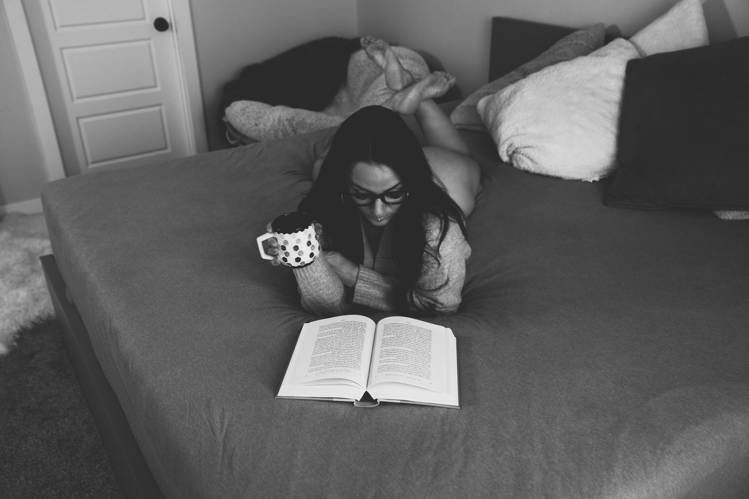 Black and white photo of woman with glasses reading book on bed while holding a mug.