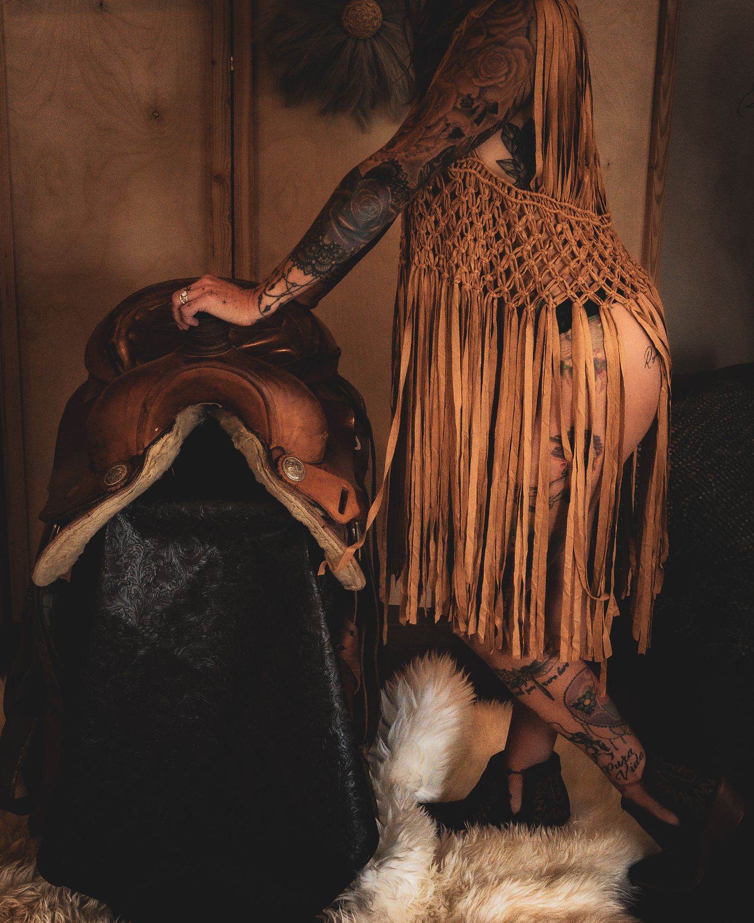 Tattooed woman in fringed suede outfit posing beside a leather saddle on a fur rug.