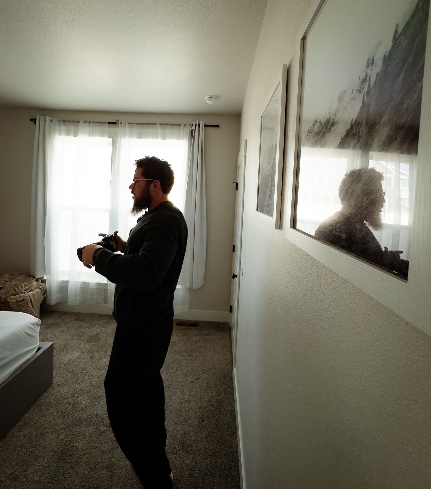 A bearded male photographer holding a camera stands in a bedroom near a window during a boudoir shoot.
