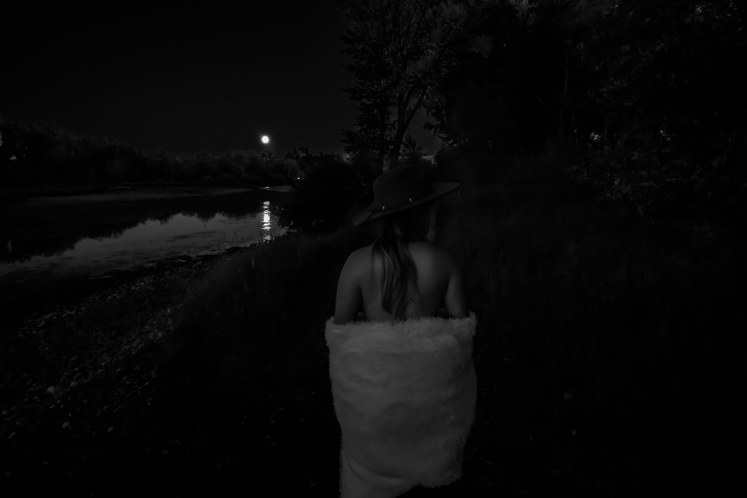 Black and white photo of a woman wrapped in a towel standing near a reflective lake at night.