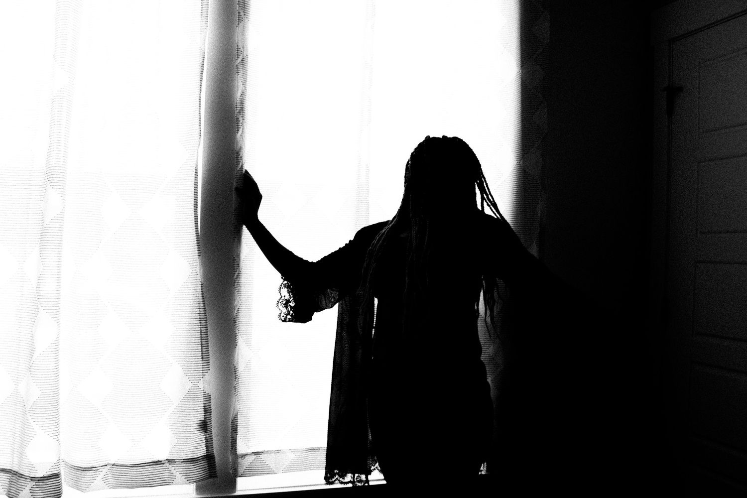 Dramatic black and white silhouette of a woman standing by sheer curtains near a bright window.