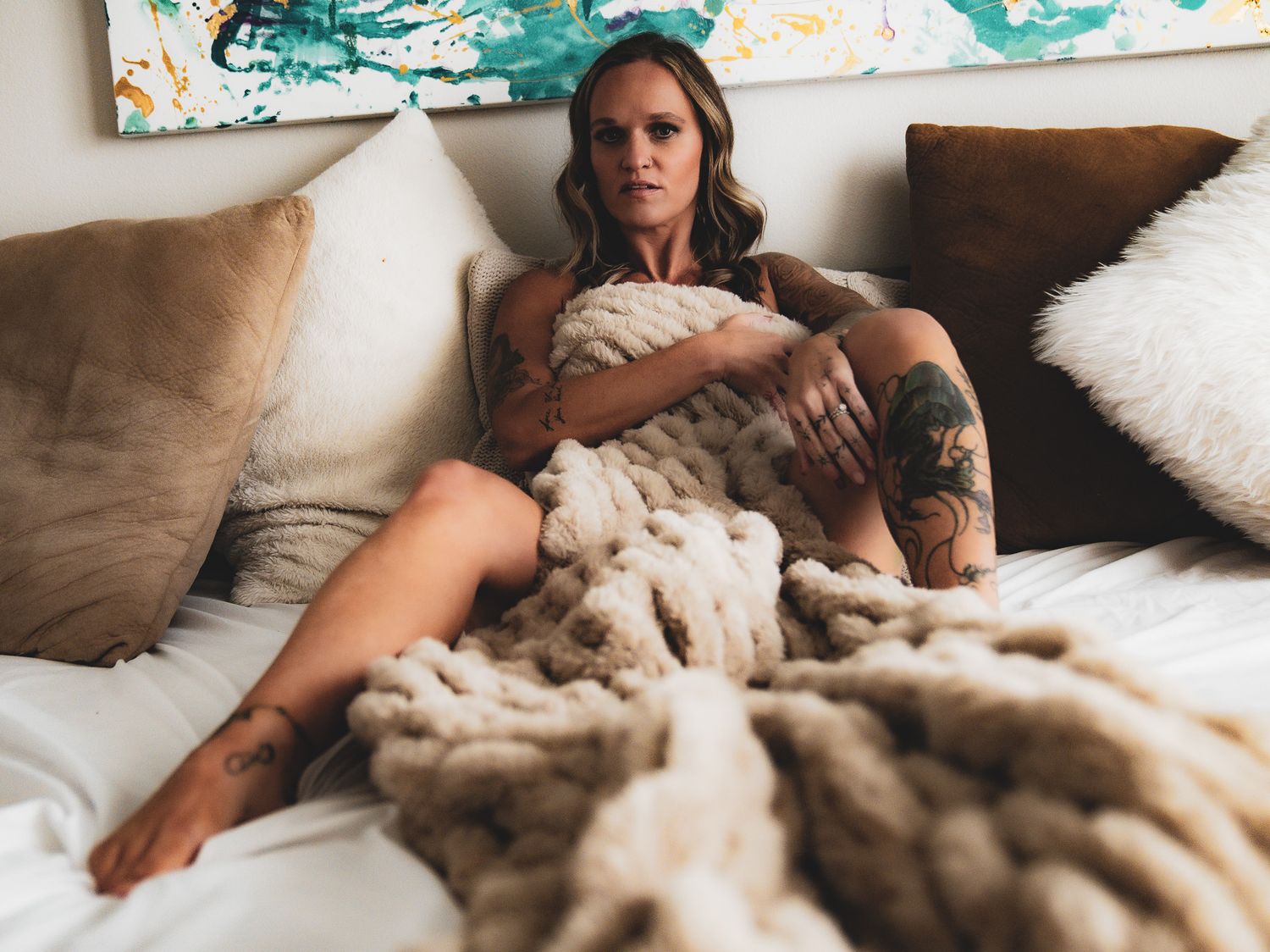 Woman wrapped in chunky knit blanket sitting on white bed with decorative pillows.