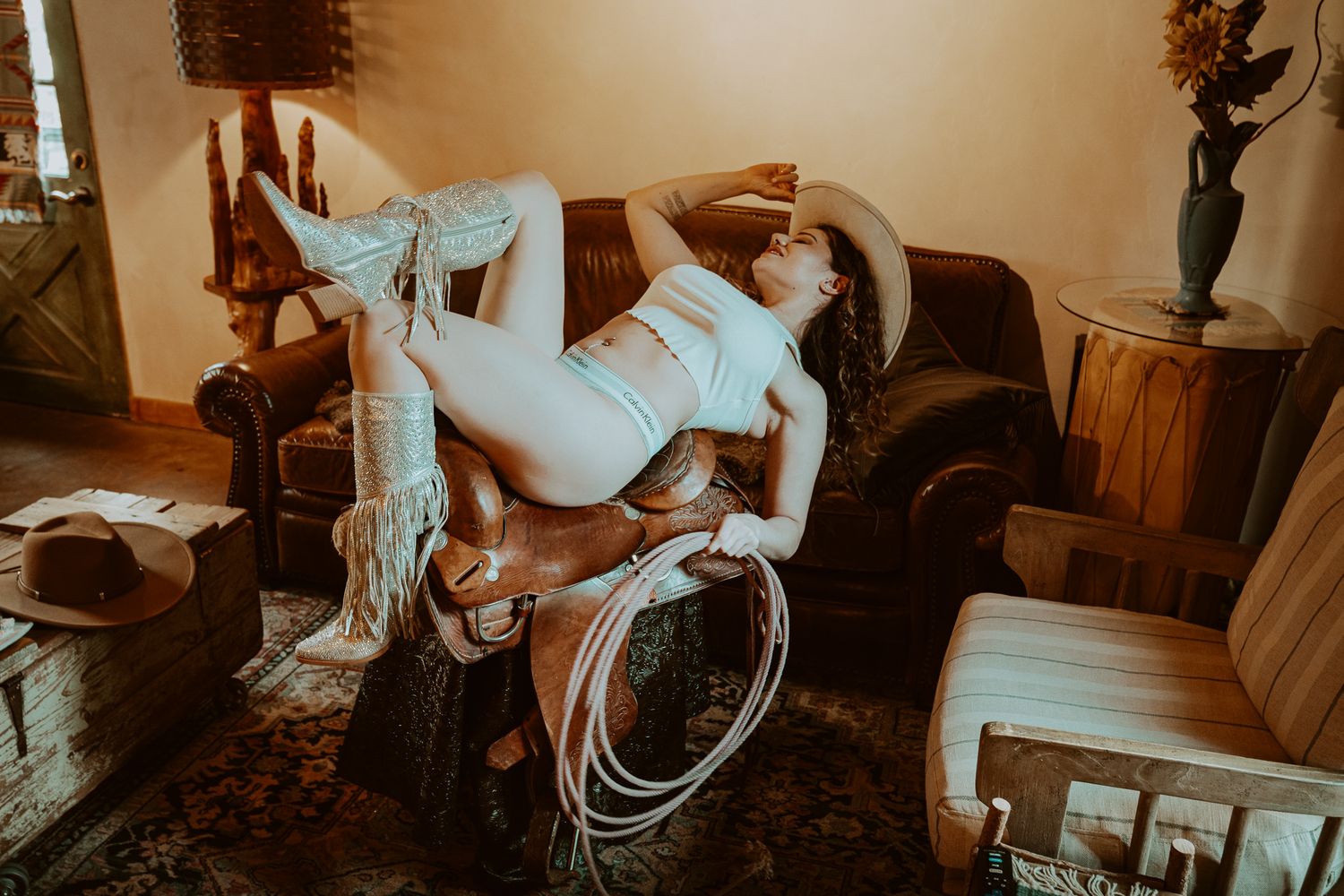 Woman in white bikini and cowboy boots posing on a saddle indoors with western-themed decor.