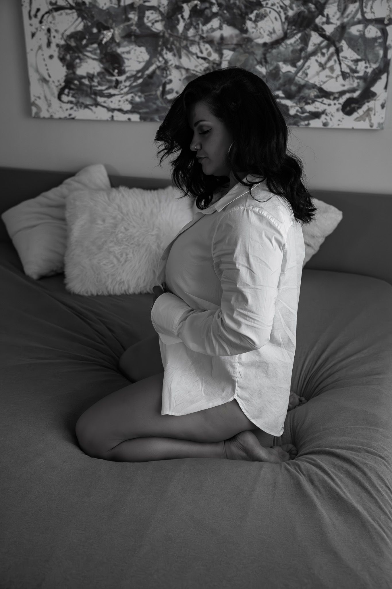 Black and white photo of brunette woman kneeling on bed in white shirt with abstract art on wall.