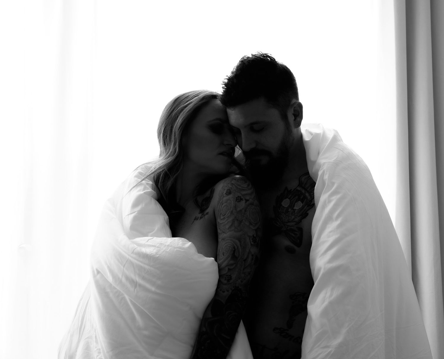 Couple wrapped in white blanket sharing intimate moment in black and white boudoir photo.