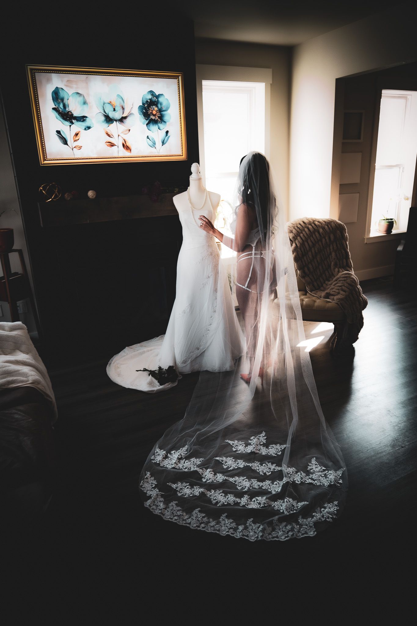 Bride in sheer lingerie viewing wedding dress on mannequin, long lace-trimmed veil trailing on floor.