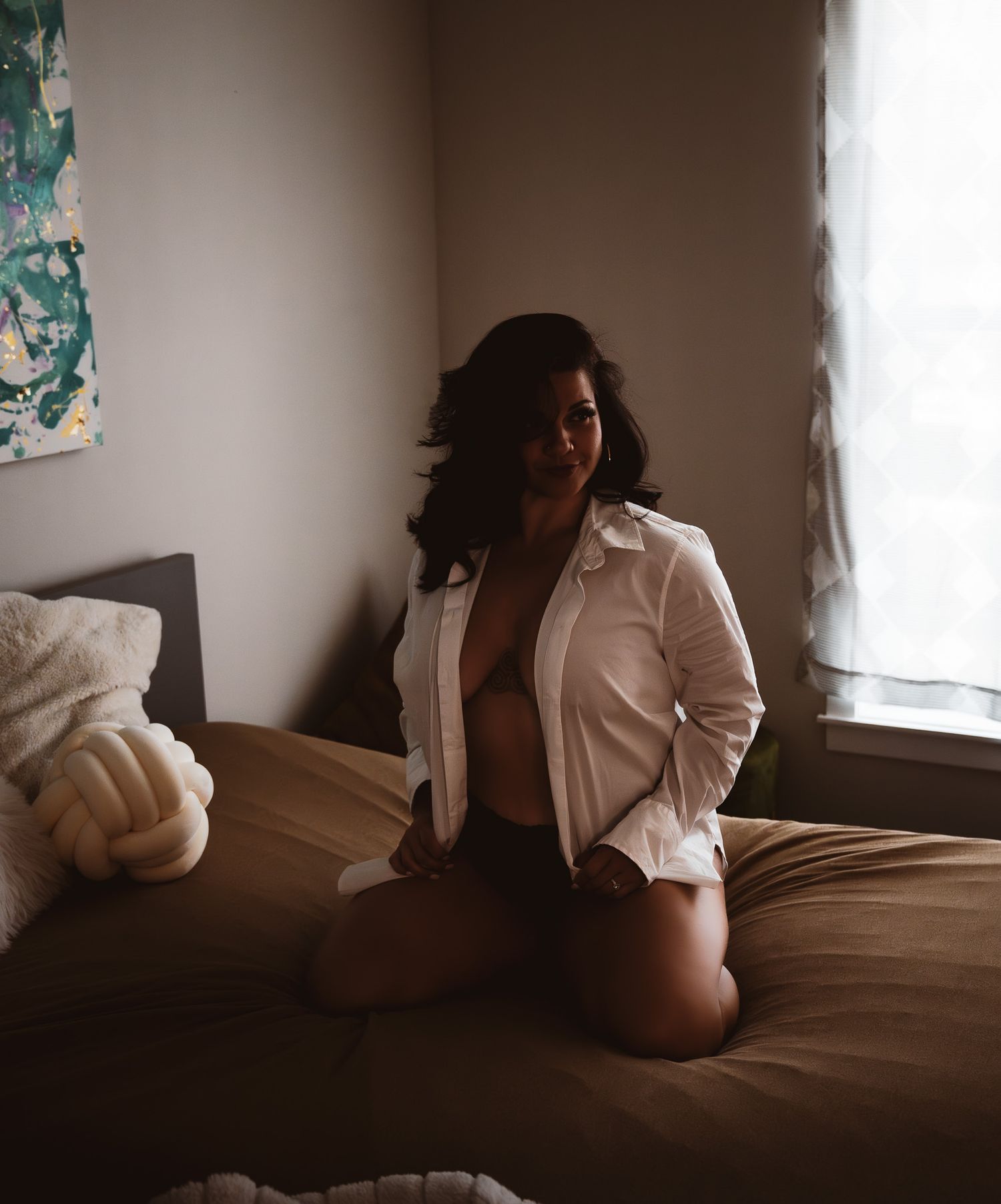 A woman in an open white shirt poses on a bed in a softly lit bedroom.