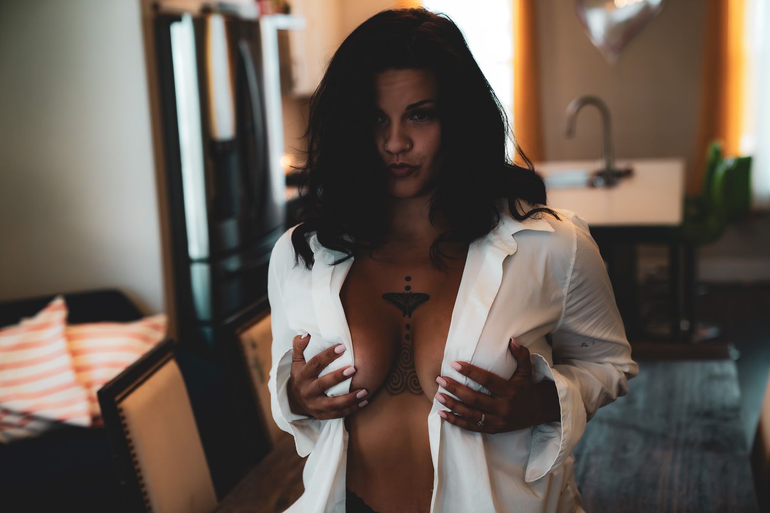 Brunette woman in open white robe with tattoos posing in dimly lit room interior.