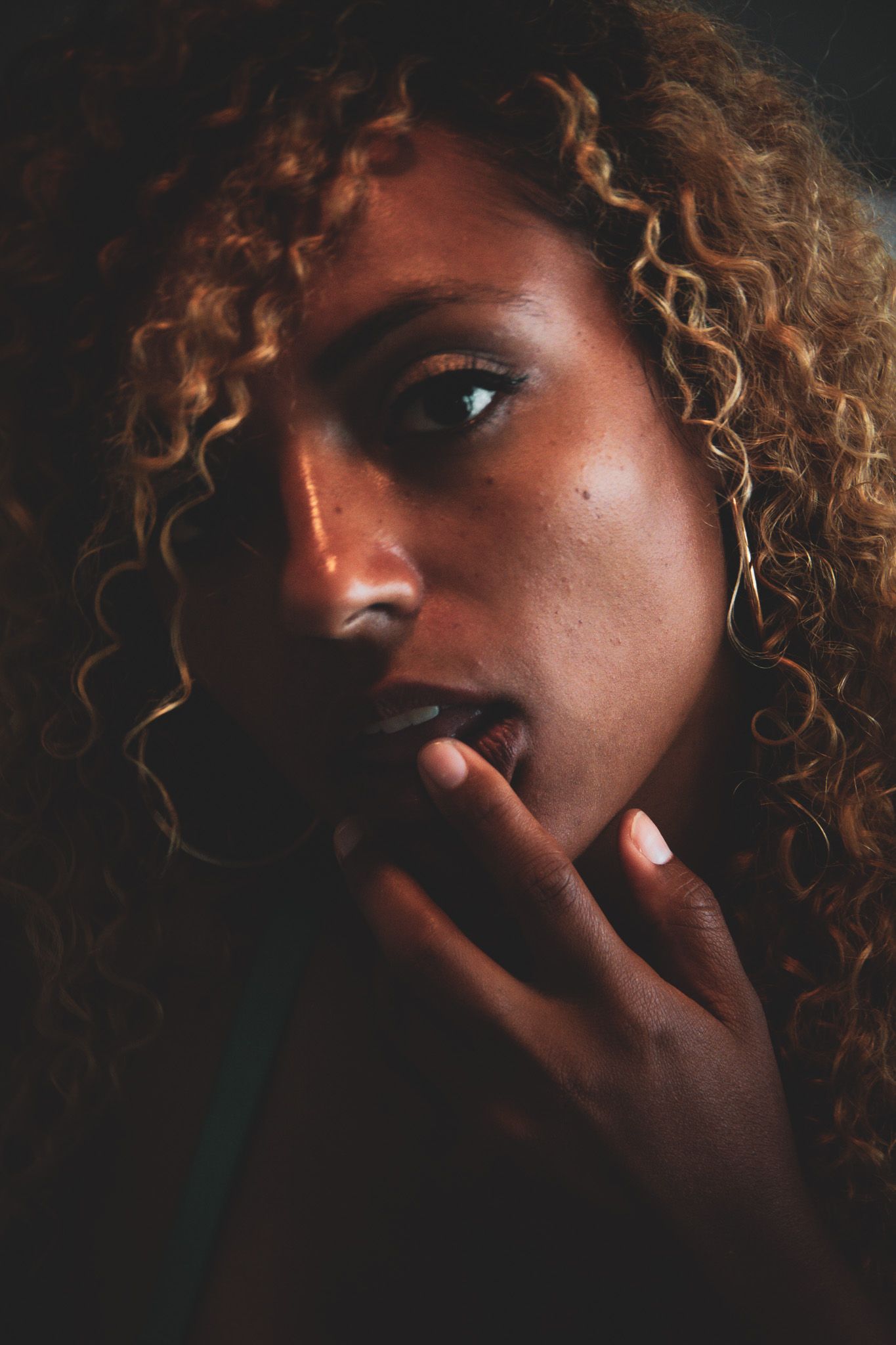 Close-up portrait of a woman with curly auburn hair in dramatic low-key lighting touching her lips.