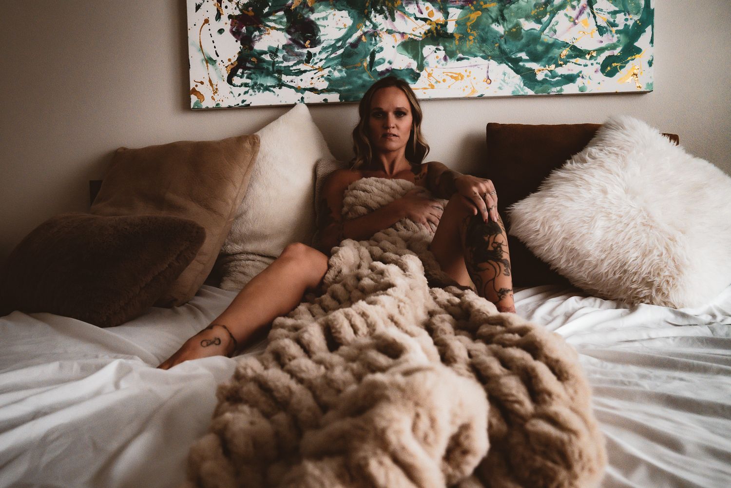 Tattooed woman relaxing on bed surrounded by pillows and chunky knit blanket in cozy bedroom.