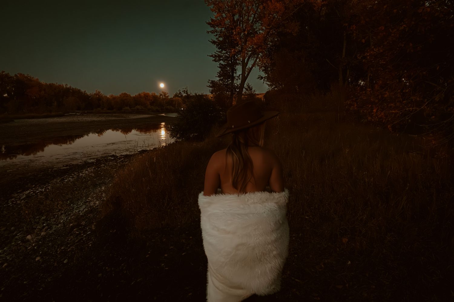 Woman wrapped in white towel standing outdoors near a moonlit lake at night among autumn trees.