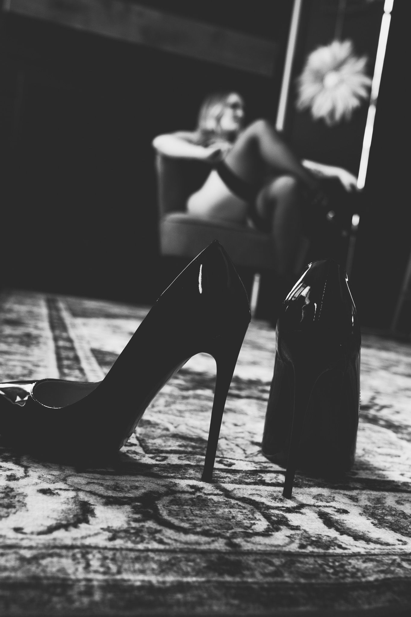 Black and white boudoir photo with high heel shoes in foreground and woman seated in background.