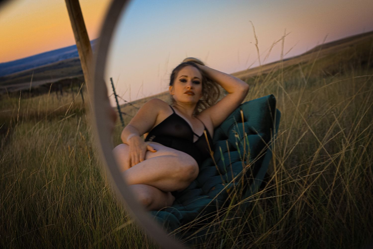 Woman in black swimsuit sitting in grass at sunset reflected in a circular mirror outdoors.