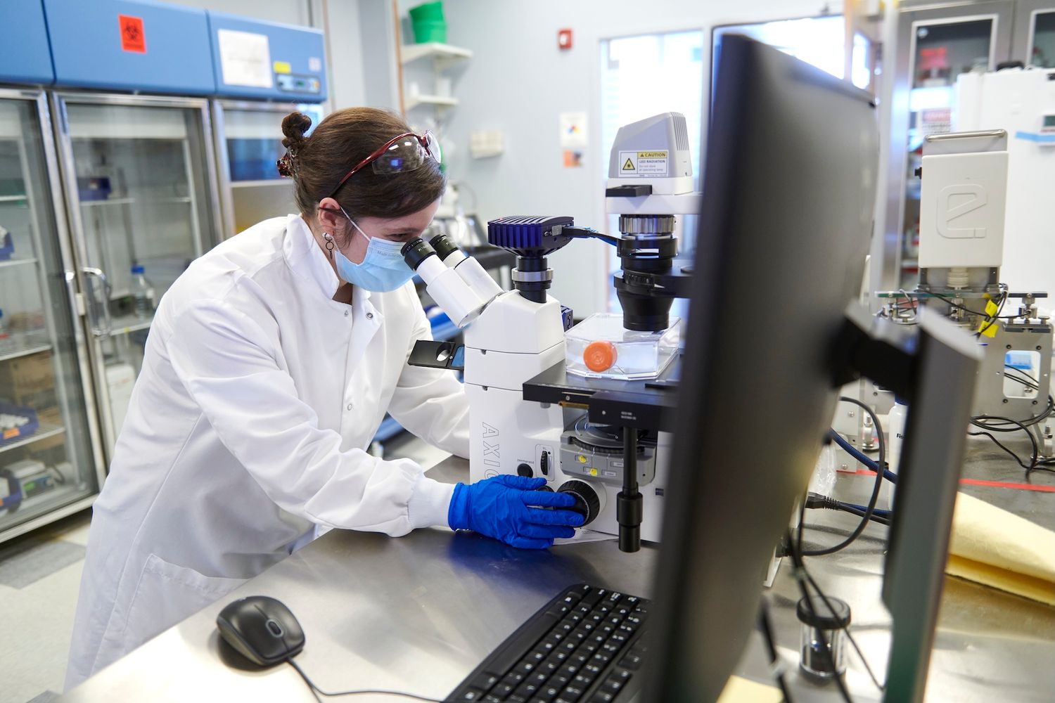 Female scientist wearing mask and safety glasses examining samples under microscope in professional lab.