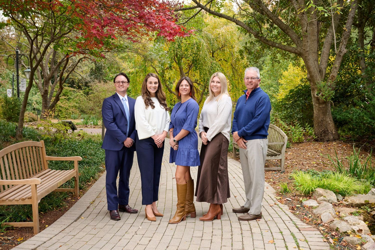 Group of five professional adults smiling together on brick garden path surrounded by autumn foliage.