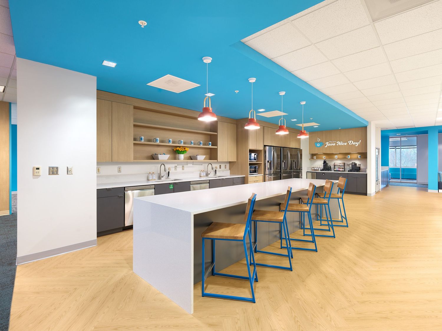 Modern office kitchen with blue ceiling, white island, bar stools, and pendant lighting.