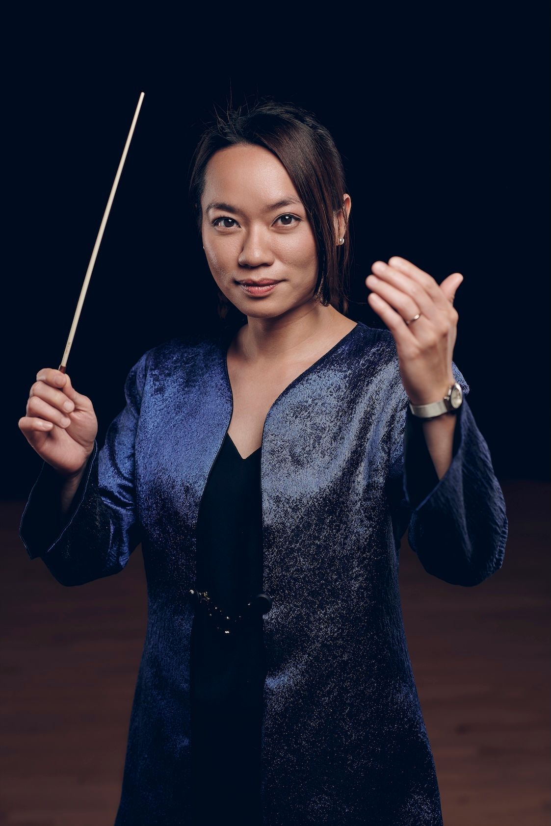 Female orchestra conductor in blue jacket holds baton dramatically against dark background.