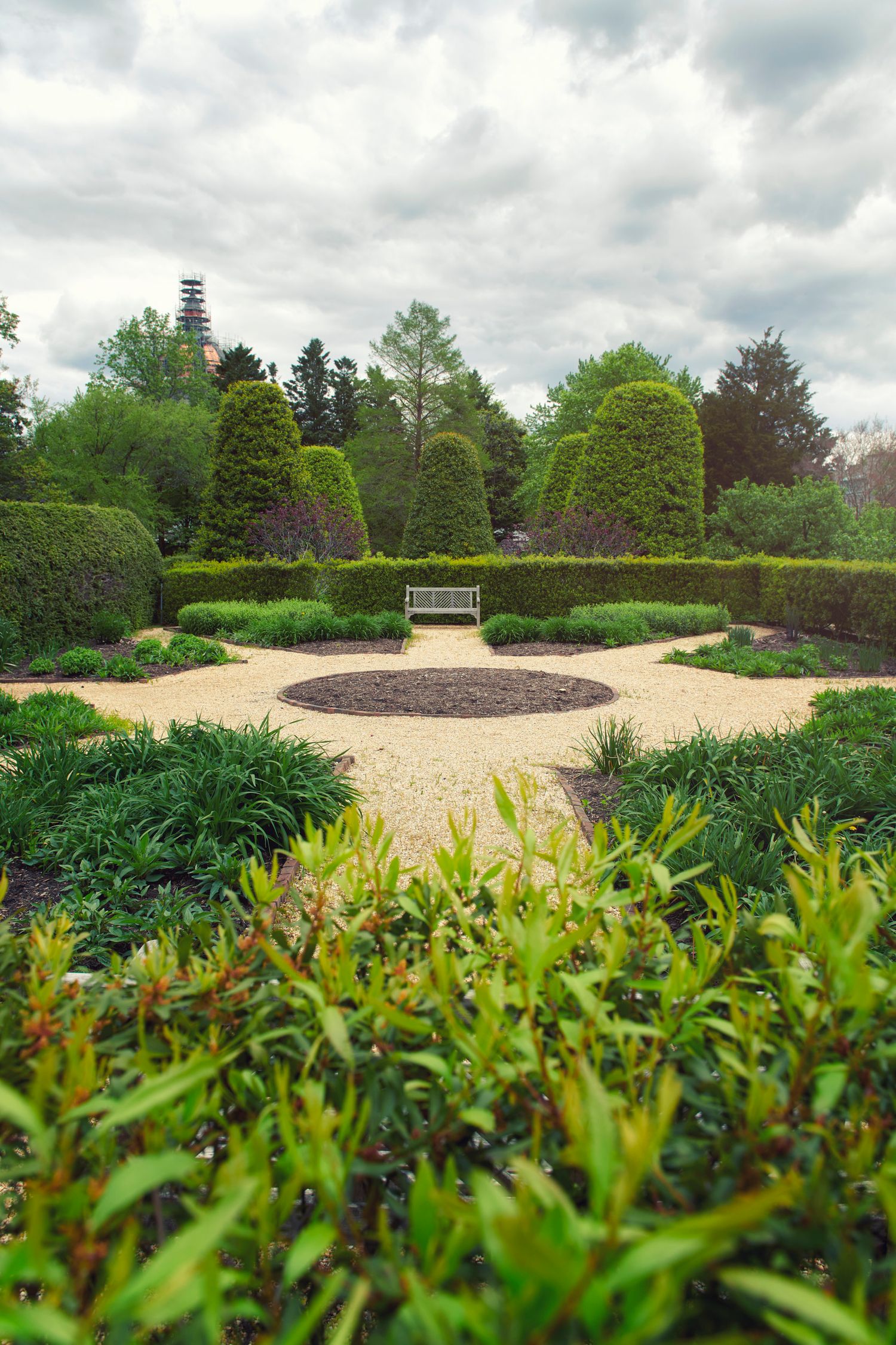 Formal English garden with manicured hedges, gravel paths, circular centerpiece, and wooden bench under cloudy sky.
