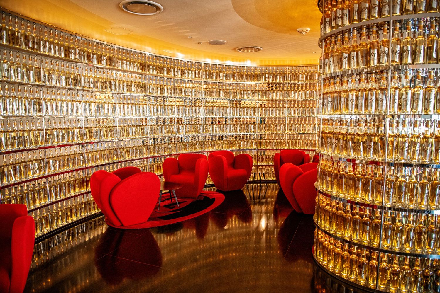Luxurious circular lounge with red velvet chairs surrounded by walls of golden bottles.