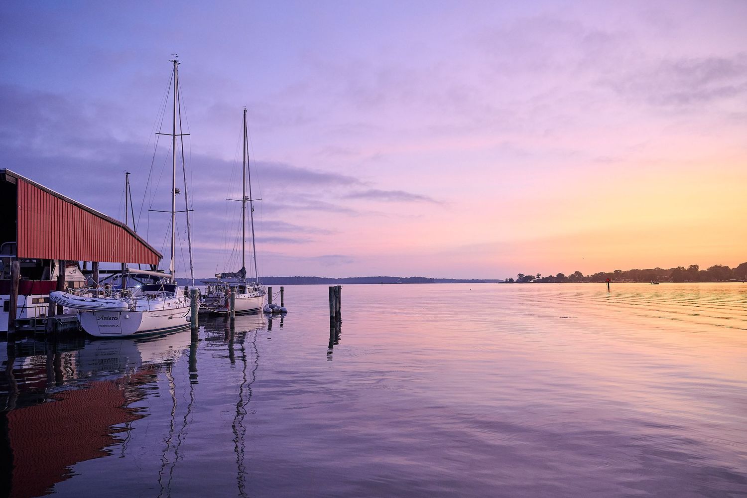 Sailboats docked at marina pier during stunning purple and orange sunset reflection.