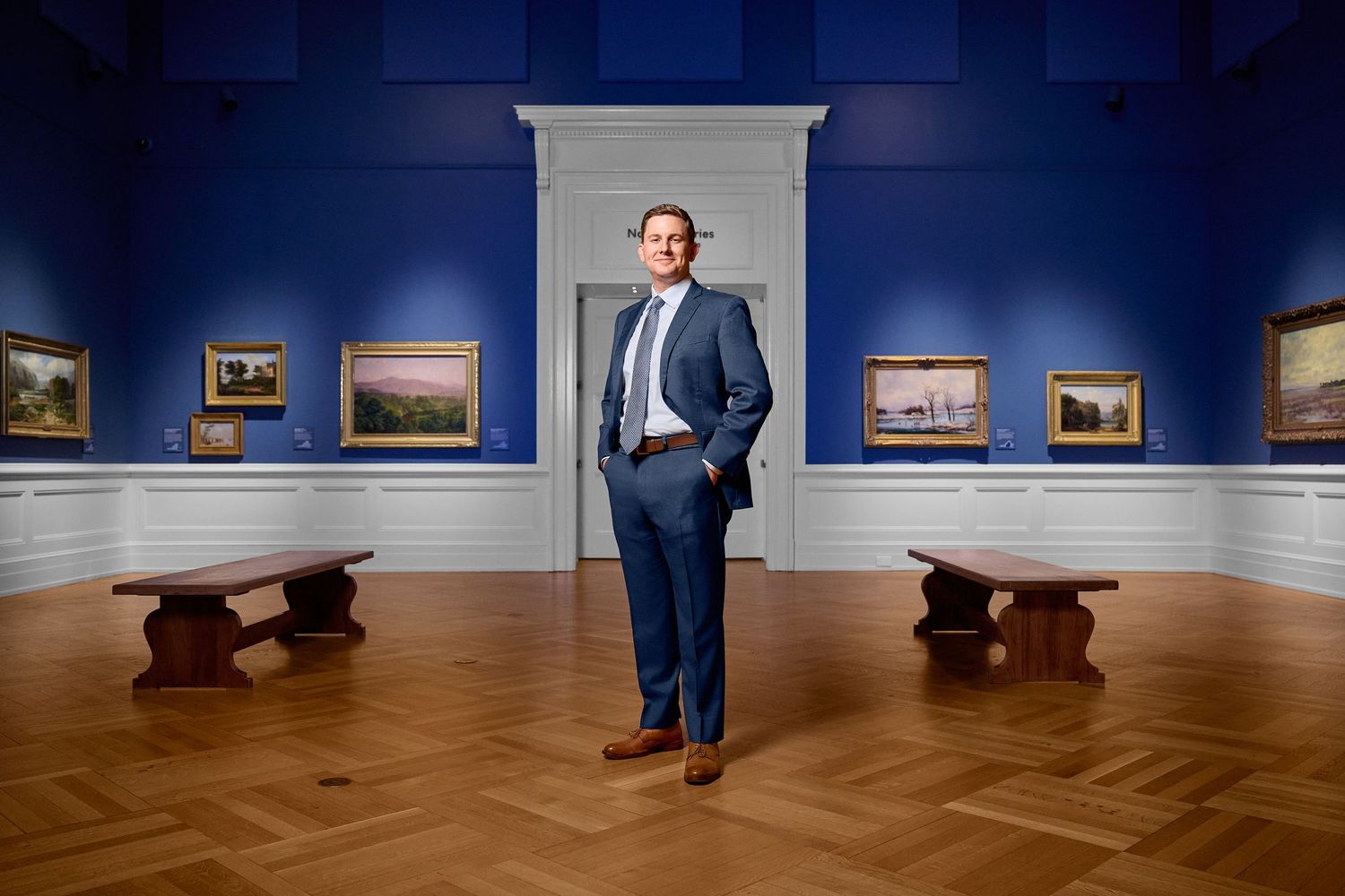 Man in blue suit standing confidently in art museum gallery with blue walls and paintings.