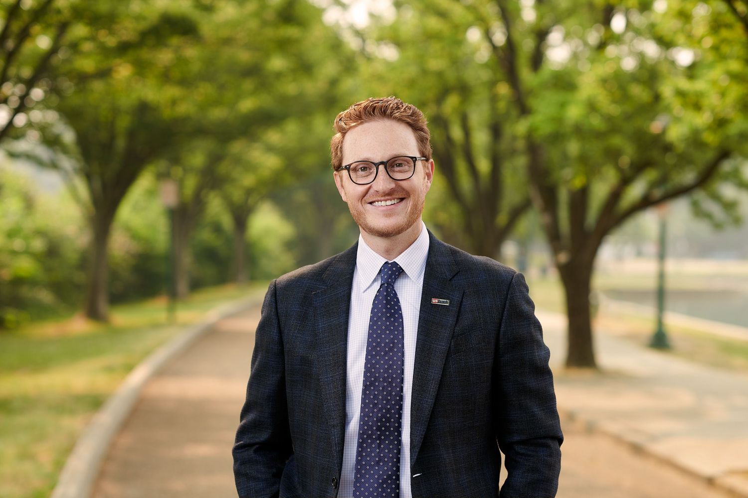 Young professional man in a navy suit smiling outdoors on a tree-lined walkway.