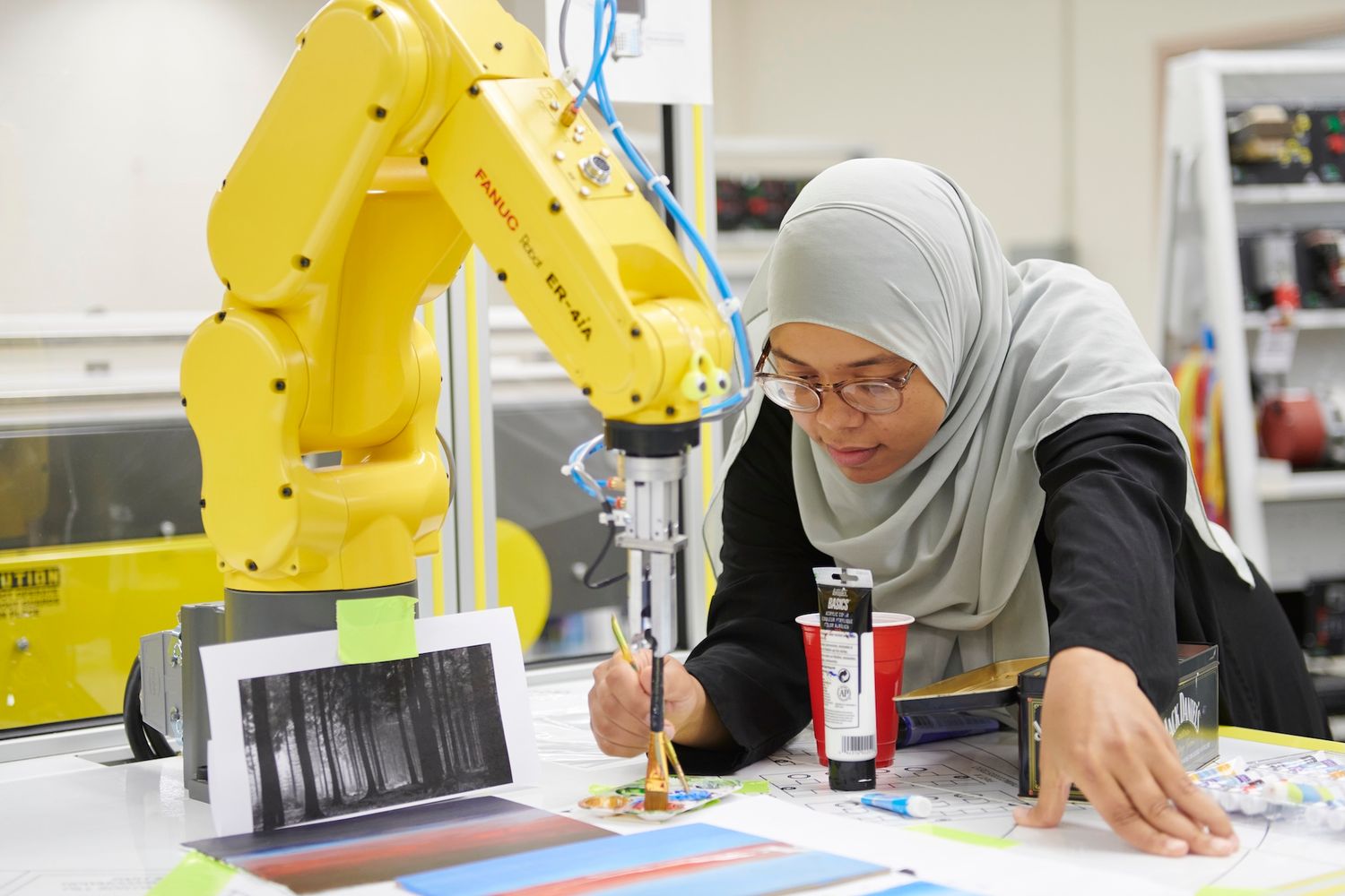 A woman in hijab works alongside a yellow robotic arm in a modern manufacturing lab.