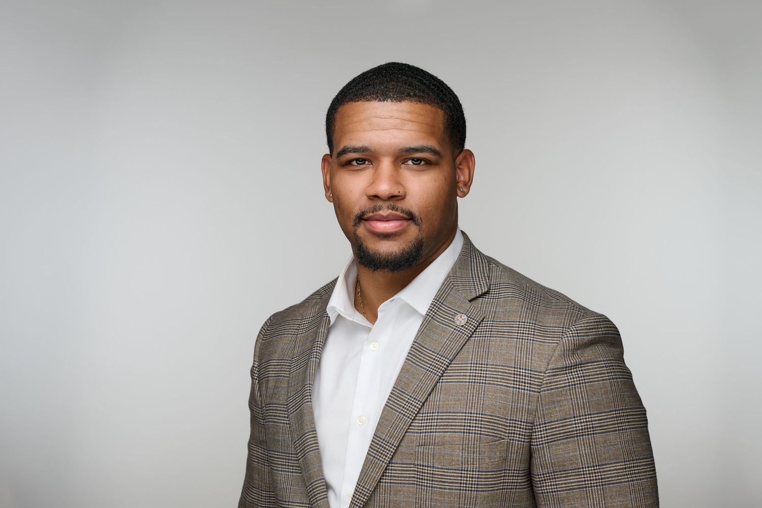 Professional headshot of confident Black man in brown plaid blazer on gray background.
