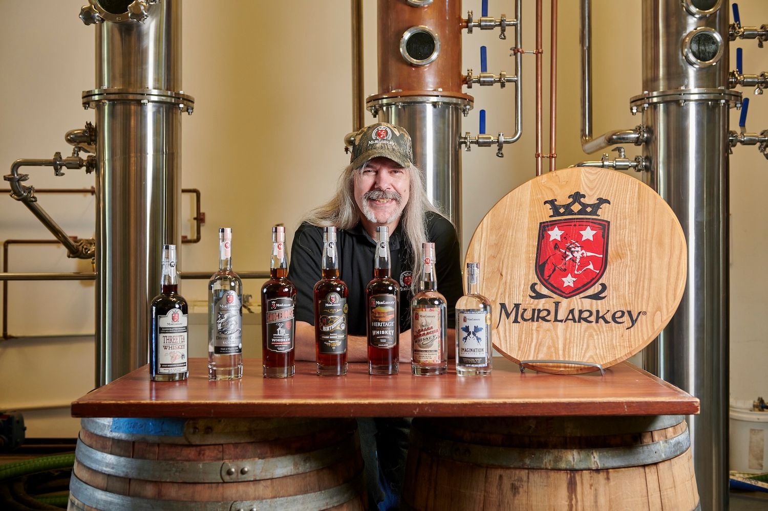 Long-haired man posing with MurLarkey whiskey bottles on barrel in front of copper distillery stills.