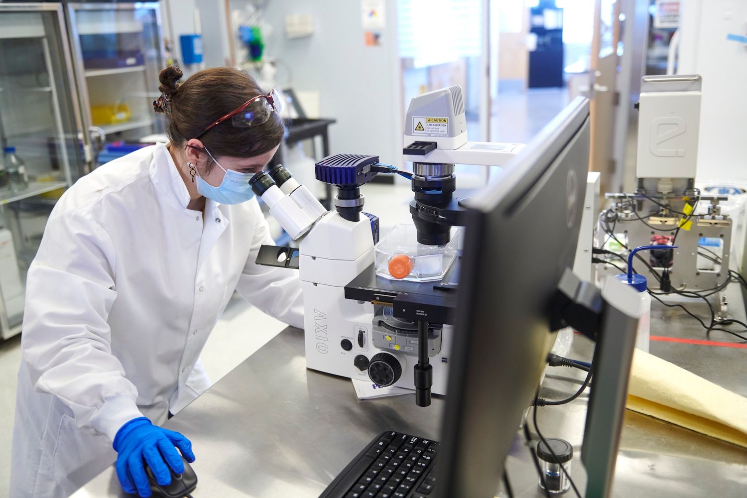 Female scientist in lab coat and safety goggles using microscope in professional research laboratory.