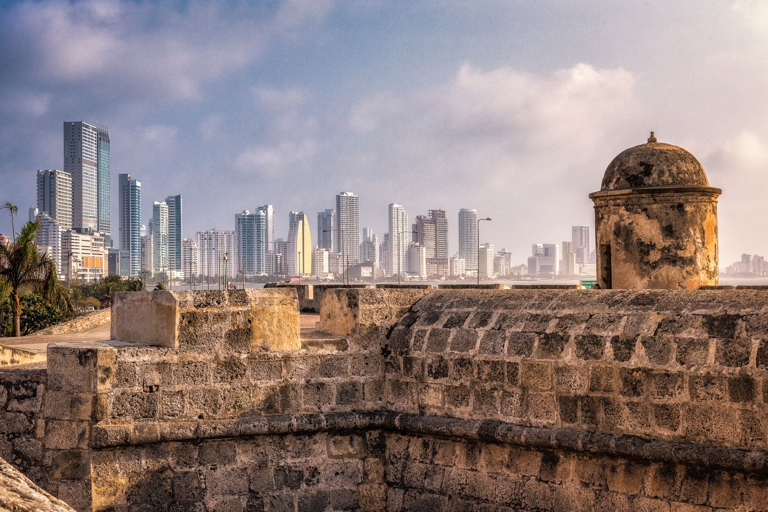 Ancient stone fortress walls with a sentry tower overlooking the modern skyline of Cartagena, Colombia.