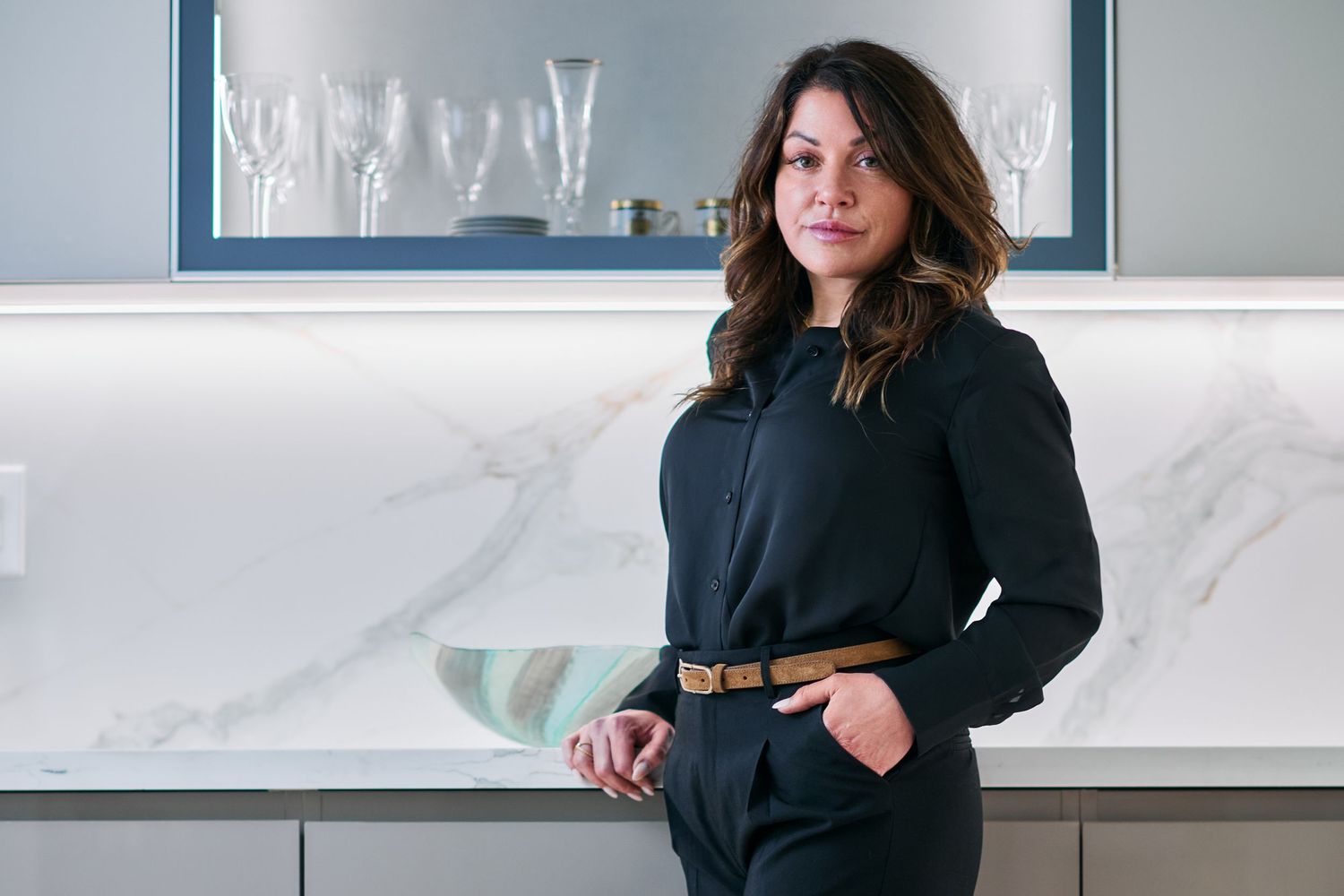 Professional woman in a black outfit with a tan belt standing in a modern kitchen interior.