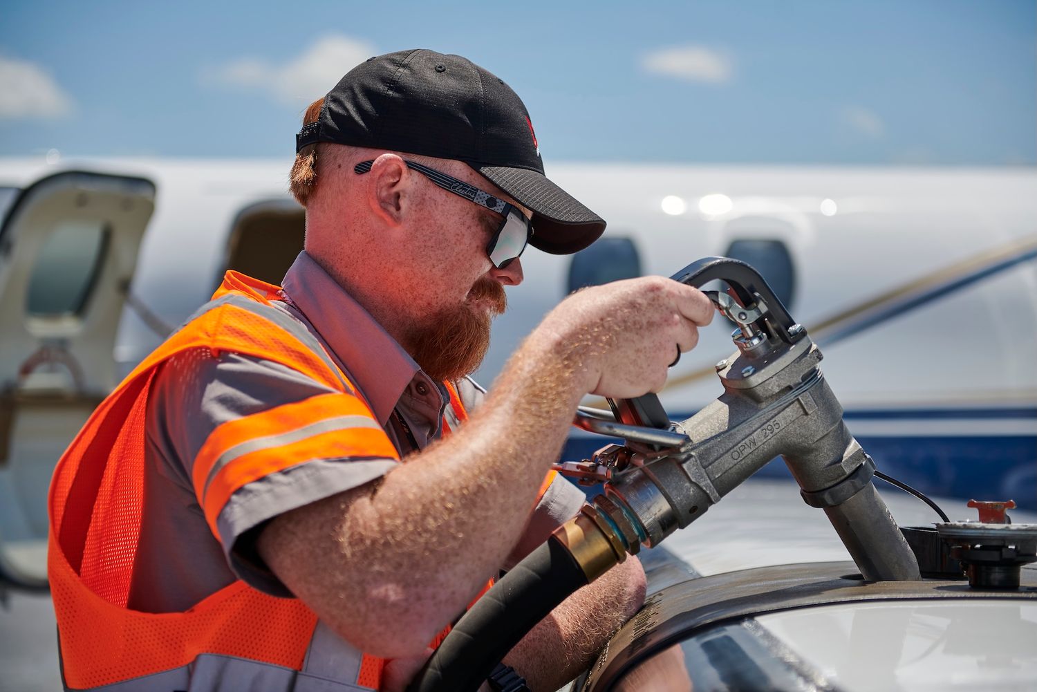 A airport ground crew worker in an orange safety vest refueling an aircraft on a sunny day.