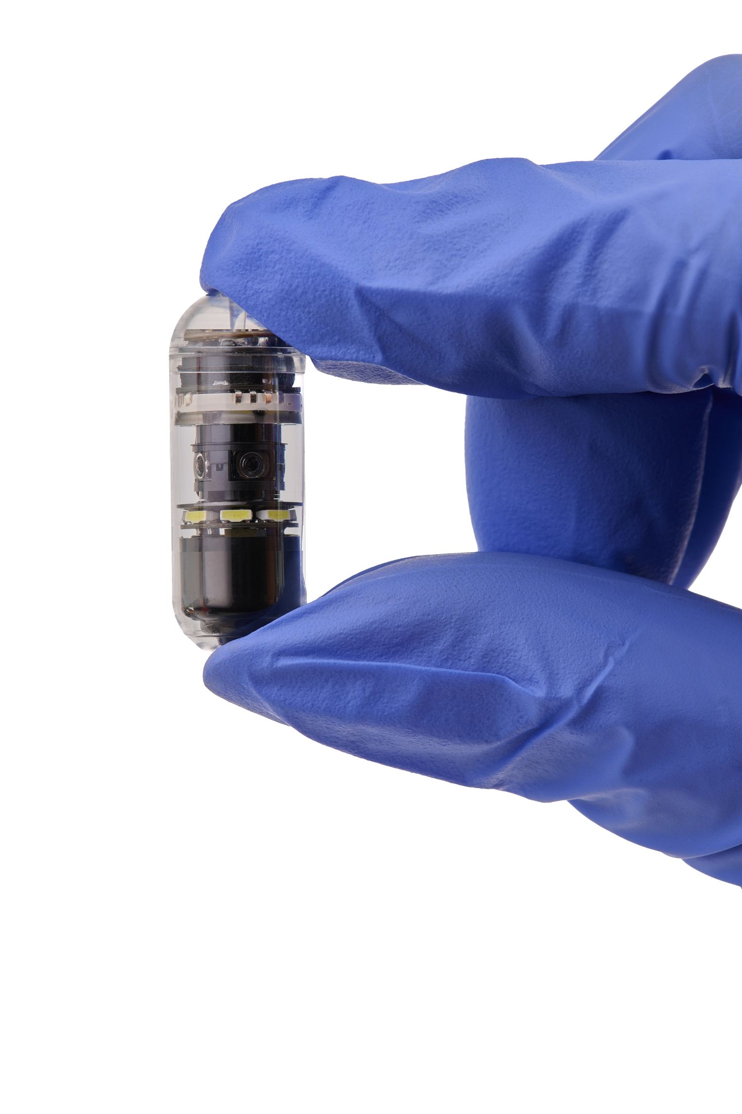 Gloved hand holding a small capsule endoscopy camera device against a white background.