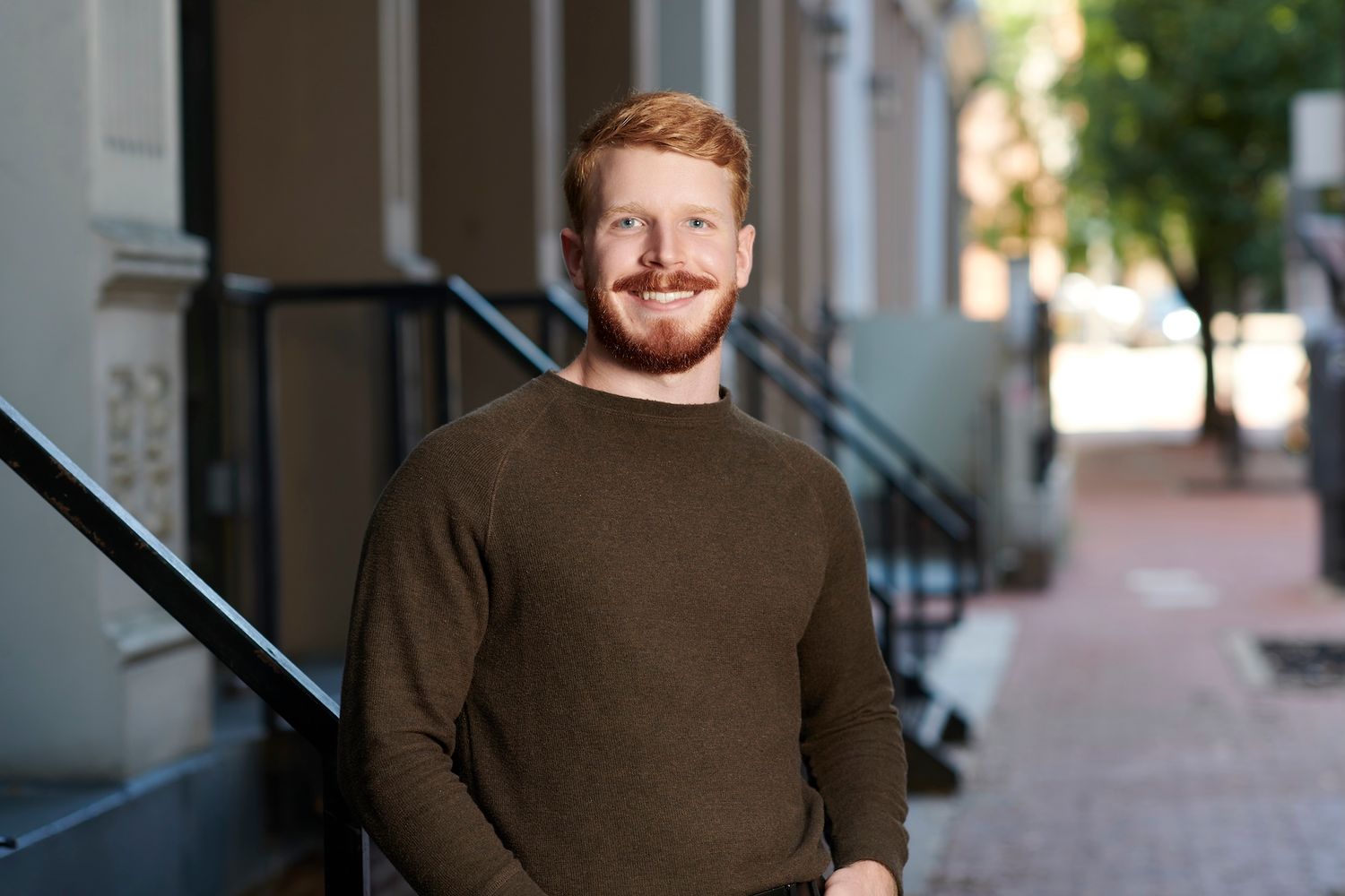 Smiling redheaded man with beard wearing olive sweater standing outdoors on a city sidewalk.